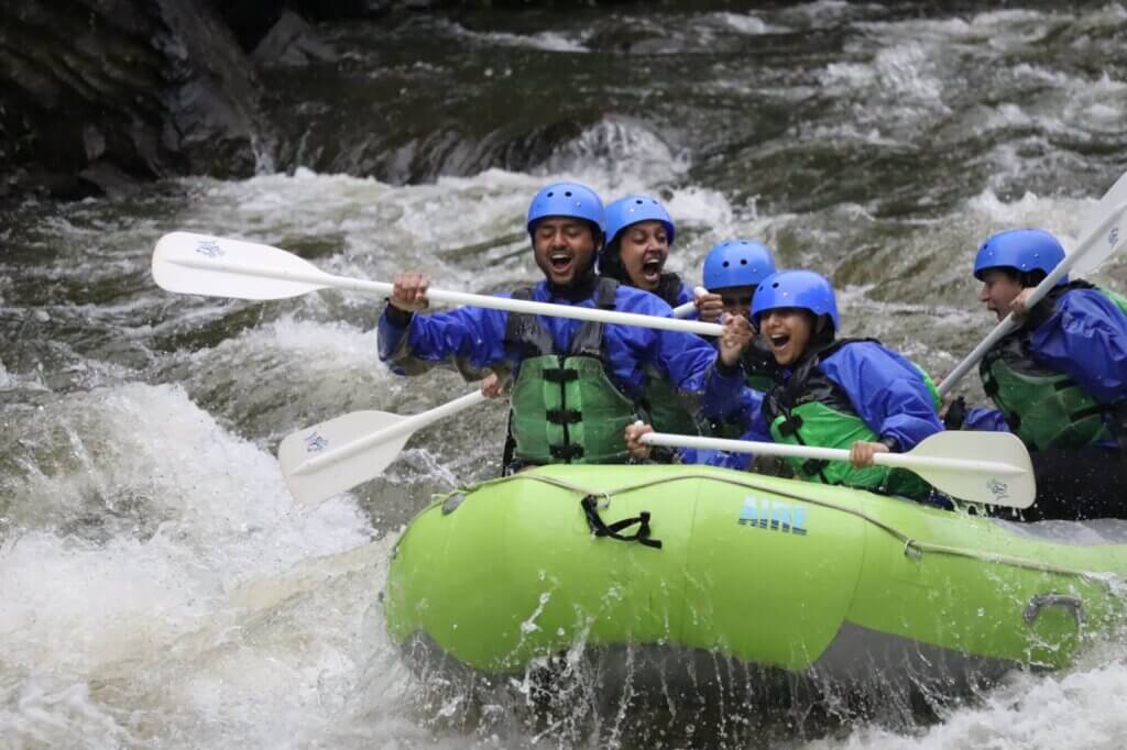 Rafters in blue helmets paddling a green inflatable raft through white water